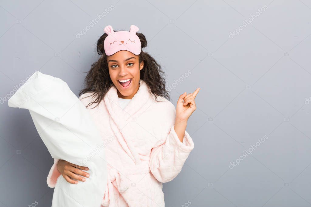 Young african american woman wearing a pajamas and a sleep mask holding a pillow smiling cheerfully pointing with forefinger away.