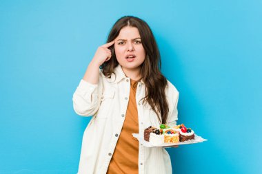Genç, kıvrımlı bir kadın elinde tatlı bir pasta tutuyor ve işaret parmağıyla hayal kırıklığı jesti yapıyor..