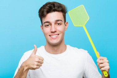 Young caucasian man holding a fly swatter smiling and raising thumb up clipart