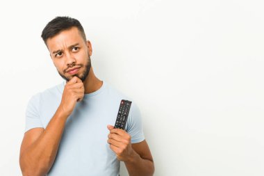 Güney Asyalı genç adam elinde bir televizyon kumandası kuşkulu ve şüpheci bir ifadeyle yana bakıyor..