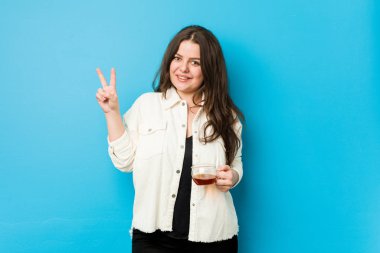 Genç kavisli kadın elinde çay fincanı tutarken neşeli ve kaygısız parmaklarıyla barış sembolü gösteriyor..