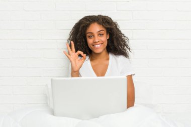 Genç Afro-Amerikan kadın dizüstü bilgisayarı ile yatakta oturuyor neşeli ve kendinden emin bir şekilde..