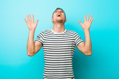 Mavi bir duvara yaslanmış genç, beyaz bir adam gökyüzüne haykırıyor, yukarı bakıyor, hayal kırıklığına uğramış..