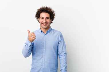 Genç, kıvırcık, olgun, zarif bir gömlek giyen, gülümseyen ve başparmağını kaldıran bir adam.