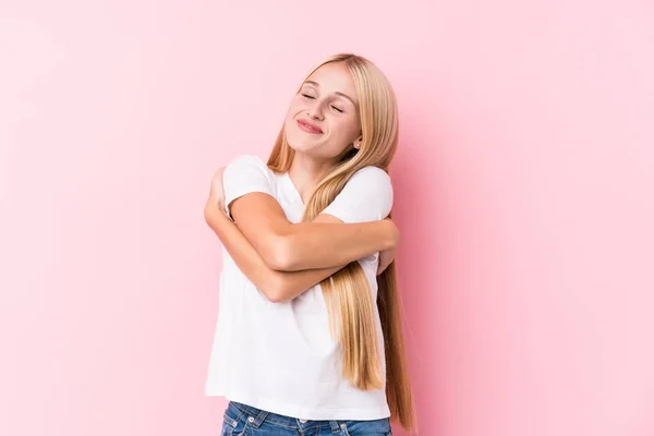 Young blonde woman on pink background hugs, smiling carefree and happy.
