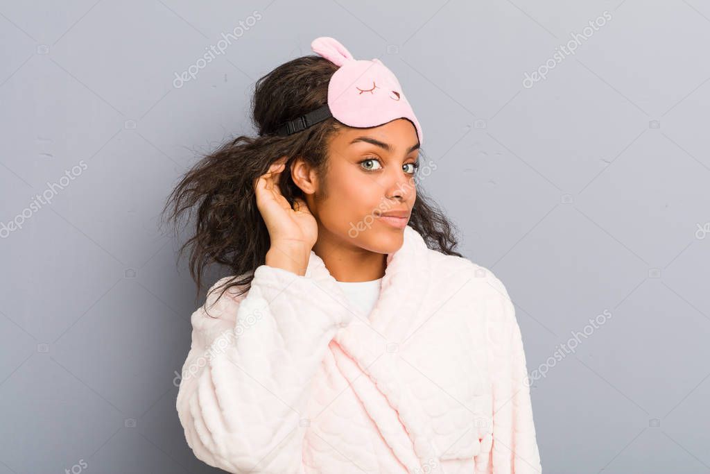 Young african american woman wearing a pajamas and a sleep mask trying to listening a gossip.