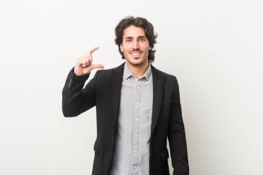 Genç bir iş adamı beyaz bir arkaplanda işaret parmaklarıyla küçük bir şey tutuyor, gülümsüyor ve kendinden emin..