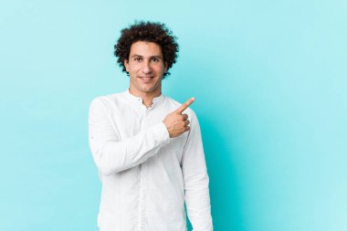 Zarif bir tişört giyen genç, kıvırcık, olgun bir adam gülümseyip, boş alanda bir şeyler gösteriyor..