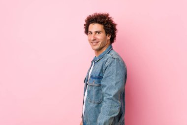 Pembe arka planda kot ceket giyen kıvırcık, olgun bir adam gülümseyen, neşeli ve keyifli görünüyor..
