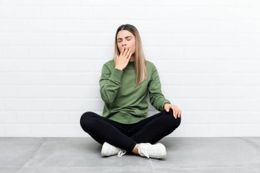 Genç beyaz kadın yerde oturuyor, esniyor. Ağzı eliyle kapatan yorgun bir hareket sergiliyor..