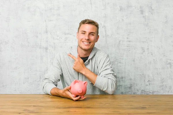Młody Człowiek Oszczędzający Pieniądze Uśmiechniętą Świnką Boku Pokazującą Coś Pustej — Zdjęcie stockowe