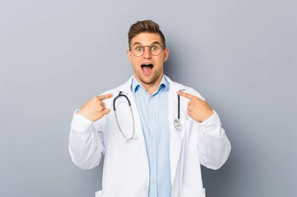 Jovem Loira Médico Homem Surpreso Apontando Para Mesmo Sorrindo Amplamente — Fotografia de Stock