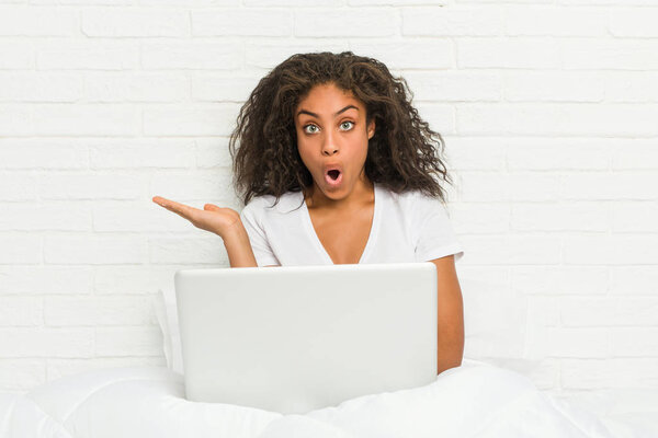 Молодая африканская американка сидит на кровати с ноутбуком впечатлен держа копировальное место на ладони
.