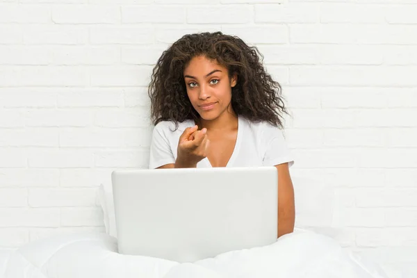 Young african american woman sitting on the bed with laptop pointing with finger at you as if inviting come closer.