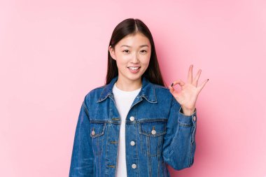 Pembe arka planda poz veren genç Çinli kadın neşeli ve kendinden emin bir şekilde iyi niyet gösteriyor..