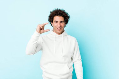 Mavi arka planda genç bir adam işaret parmaklarıyla küçük bir şey tutuyor, gülümsüyor ve kendinden emin..