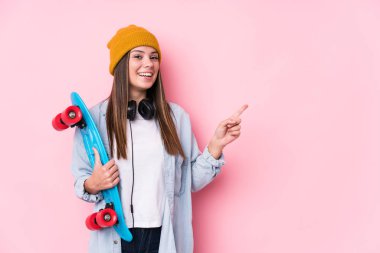 Genç patenci kadın elinde bir kaykayla gülümsüyor ve bir kenara işaret ediyor, boş alanda bir şey gösteriyor..