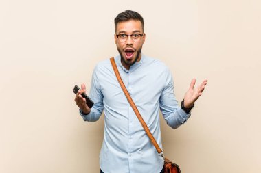 Genç melez Asyalı iş adamı elinde bir telefonla zaferi ya da başarıyı kutluyor.