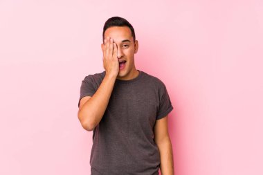 Pembe arka planda poz veren yog latin adam yüzünün yarısını avuç içiyle örterek eğleniyor..