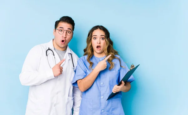 一对年轻的医生夫妇站在蓝色的背景上 孤立无援地指向一边 — 图库照片