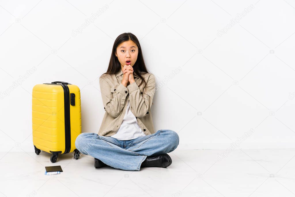 Young chinese traveler woman sitting holding a boarding passes praying for luck, amazed and opening mouth looking to front.