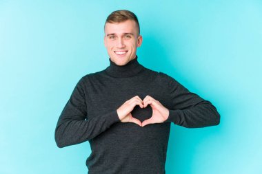 Mavi arka planda gülümseyen ve elleriyle kalp şeklini gösteren genç beyaz bir adam..