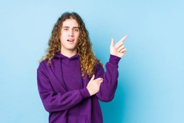 Uzun saçlı adam yalnız poz veriyor. Gülümsüyor, işaret parmağıyla işaret ediyor..