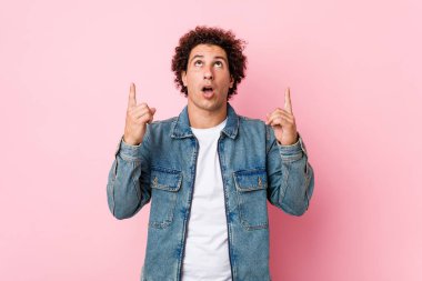 Kıvırcık, olgun bir adam pembe arka planda kot ceket giyiyor üst tarafı açık ağızla işaret ediyor..