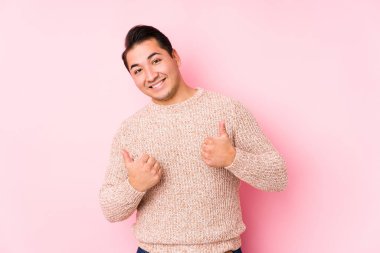 Pembe arka planda poz veren kıvrımlı genç adam iki parmağını da kaldırıyor, gülümsüyor ve kendinden emin..