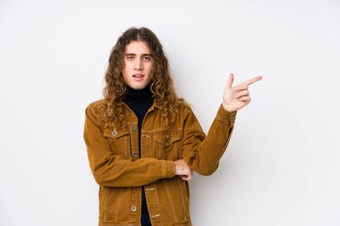 Uzun saçlı adam yalnız poz veriyor. Gülümsüyor, işaret parmağıyla işaret ediyor..