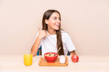 Genç, beyaz bir kadın kahvaltı yapıyor, izole edilmiş, parmaklarıyla bir cep telefonu hareketi gösteriyor..