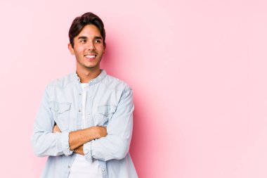 Pembe arka planda poz veren beyaz bir genç yalnız ve kollarını kavuşturarak gülümsüyor..