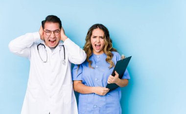 Mavi arka planda poz veren genç doktor çift kulaklarını elleriyle kapatıyor ve çok fazla ses duymamaya çalışıyorlar..