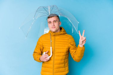 Şemsiye tutan genç beyaz adam neşeli ve kaygısız parmaklarıyla barış sembolü gösteriyor..