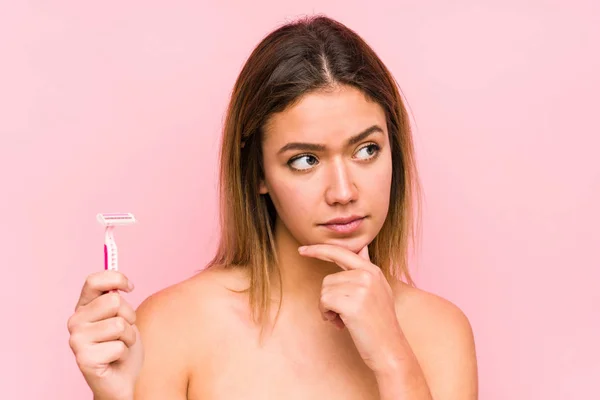 Young caucasian woman holding a razor blade isolated Young caucasian woman holding a hairbrush isolated looking sideways with doubtful and skeptical expression.