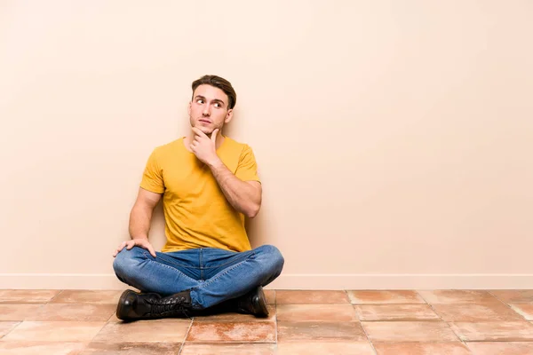 Νεαρός Καυκάσιος Που Κάθεται Στο Πάτωμα Απομονωμένος Κοιτάζοντας Πλάγια Αμφίβολη — Φωτογραφία Αρχείου