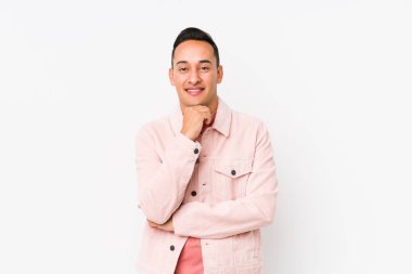 Genç latin adam yalnız poz veriyor gülümsüyor ve kendinden emin, elleriyle çeneye dokunuyor..