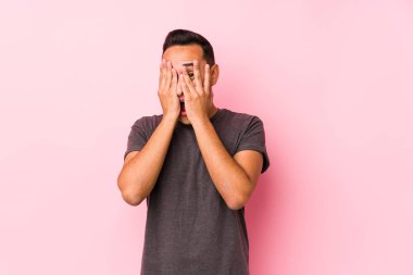 Pembe arka planda poz veren yog latin adam, parmaklarının arasından, korkmuş ve gergin bir şekilde göz kırpıyor..