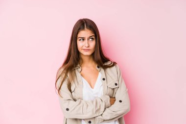 Pembe arka planda poz veren genç beyaz kadın kameraya alaycı bir ifadeyle bakıyor..