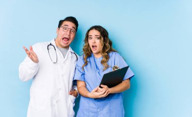 Mavi arka planda poz veren genç doktor bir zaferi ya da başarıyı kutluyor, şaşırıyor ve şok oluyor..