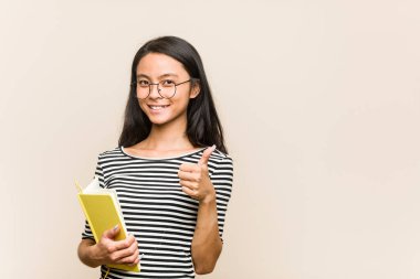 Asyalı genç bir kız öğrenci elinde bir kitap tutuyor gülümsüyor ve başparmağını kaldırıyor.