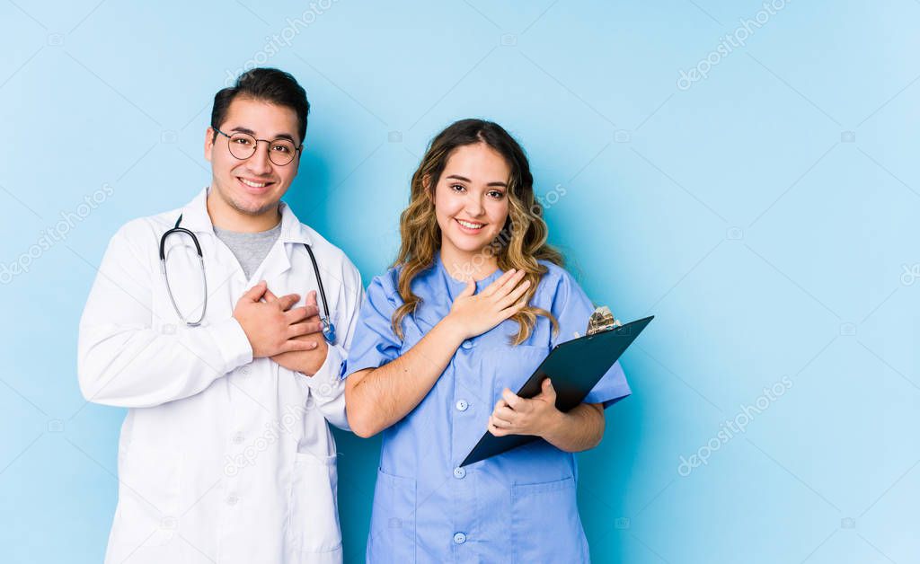 Young doctor couple posing in a blue background isolated has friendly expression, pressing palm to chest. Love concept.