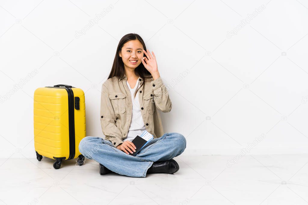 Young chinese traveler woman sitting holding a boarding passes cheerful and confident showing ok gesture.