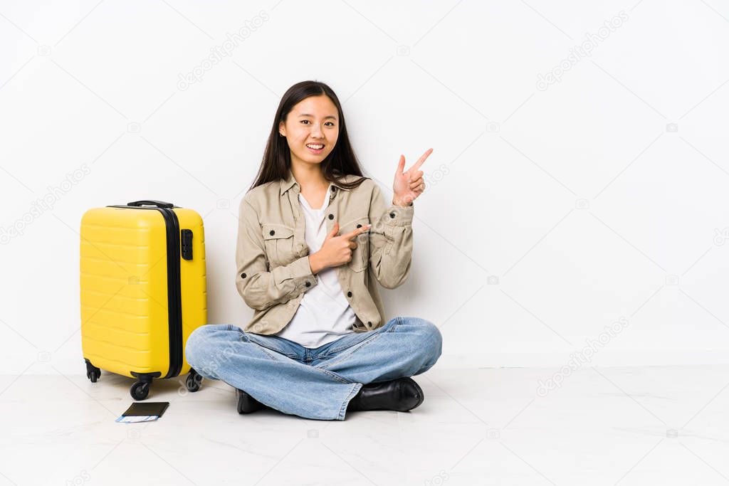 Young chinese traveler woman sitting holding a boarding passes pointing with forefingers to a copy space, expressing excitement and desire.