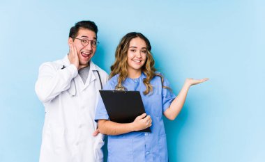 Mavi arka planda poz veren genç doktor çift avuç içlerinde fotokopi alanı tutuyor, el üstünde tutuyorlar. Şaşırmış ve mutlu..