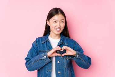 Pembe arka planda poz veren genç Çinli kadın izole edilmiş, gülümseyen ve elleriyle kalp şeklini gösteren..