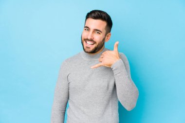 Mavi arka planda genç beyaz bir adam izole edilmiş, parmakları olan bir cep telefonu hareketi gösteriyor..