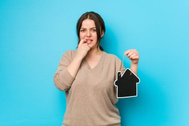 Genç ve kıvrımlı bir kadın elinde ev ikonu tutuyor tırnaklarını yiyor, gergin ve çok endişeli..