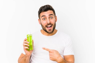 Genç, beyaz bir adam, elinde aloe vera ile nemlendirici tutarken izole edilmiş bir şekilde gülümseyerek işaret parmağıyla işaret ediyor..