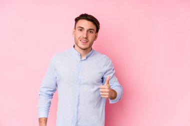 Pembe arka planda poz veren beyaz bir genç yalnız, gülümseyen ve başparmağını kaldıran.
